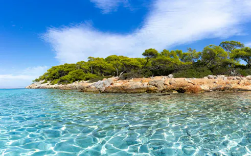 Clear waters and rocky shoreline of Moni Island, Greece, with lush greenery in the background.