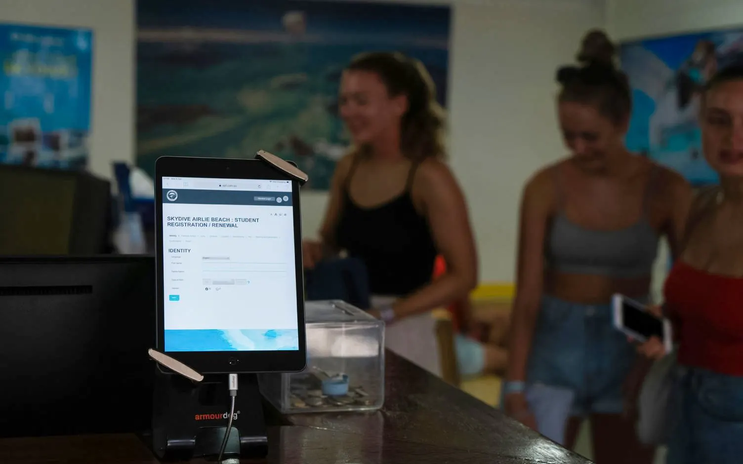 Tablet displaying skydive registration at Mission Beach with people in the background.
