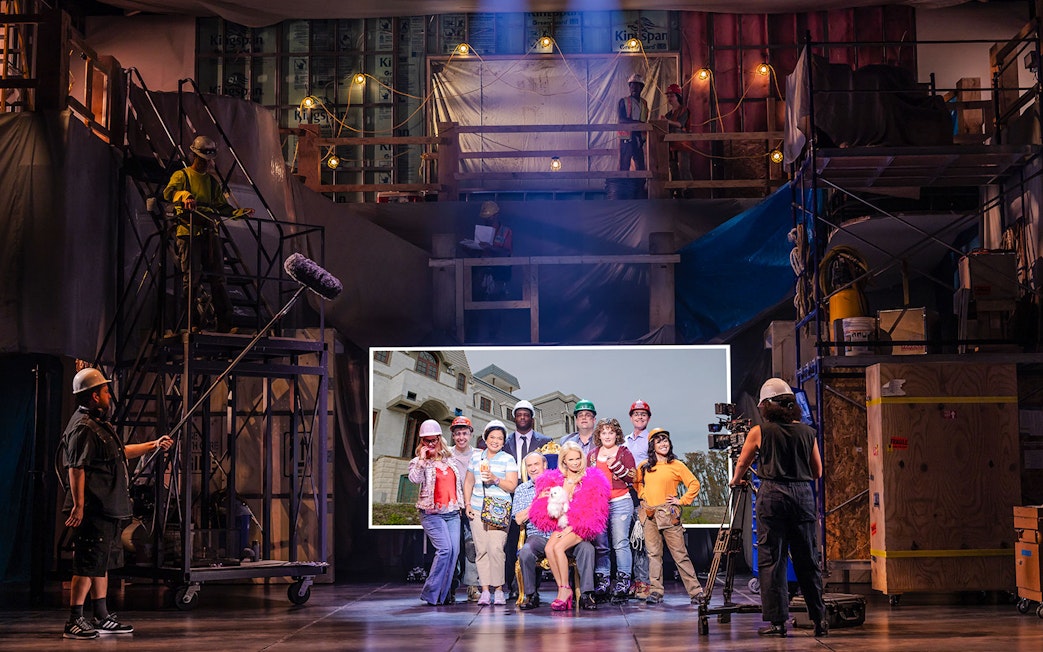 Group of people in construction gear on a stage set, with a backdrop of a mansion, related to "The Queen of Versailles.