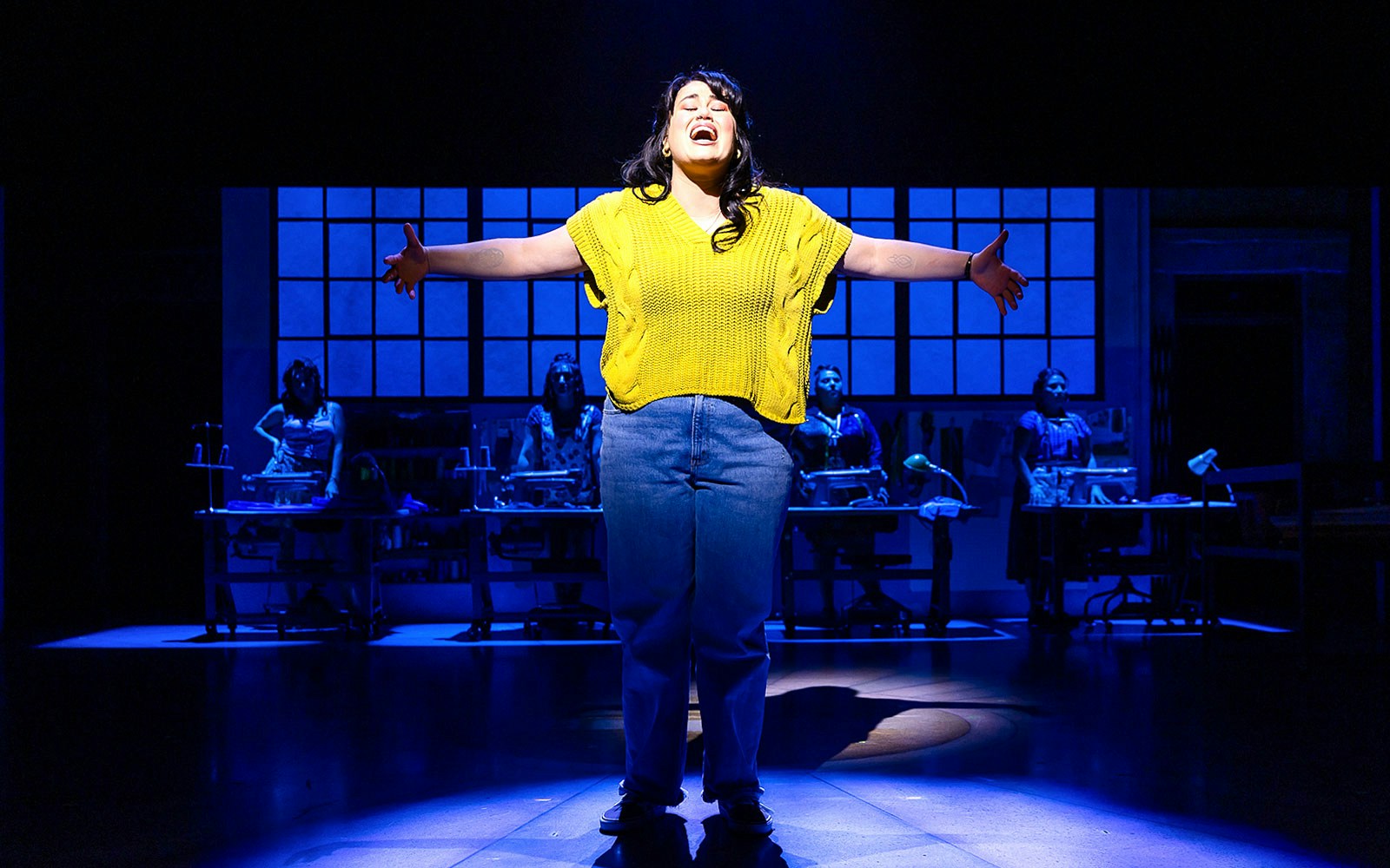 Performer singing on stage in "Real Women Have Curves: The Musical" with sewing machines in the background.