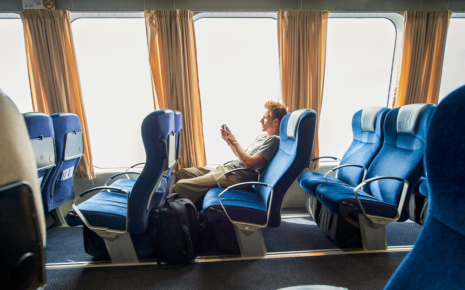 Ferry passenger seated comfortably, using a phone, with blue seats and large windows.