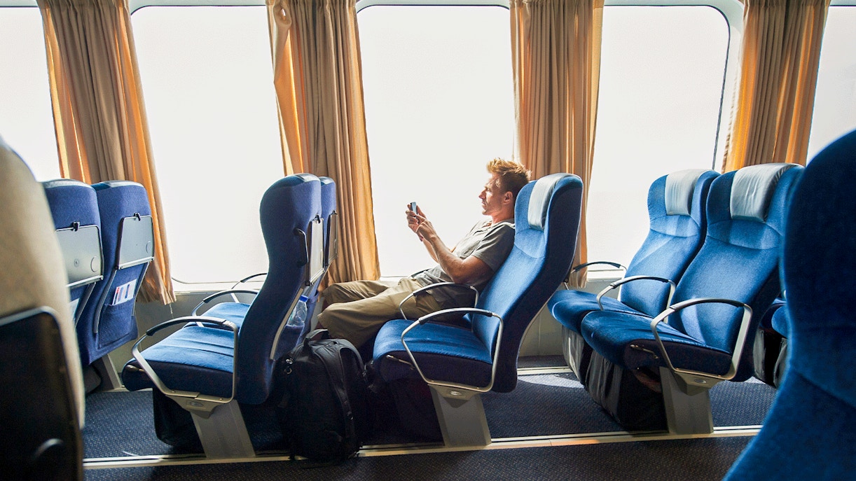 Ferry passenger seated comfortably, using a phone, with blue seats and large windows.