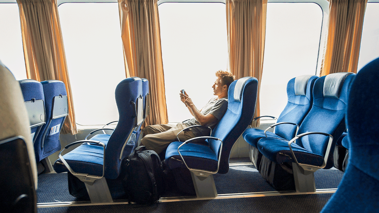 Ferry passenger seated comfortably, using a phone, with blue seats and large windows.