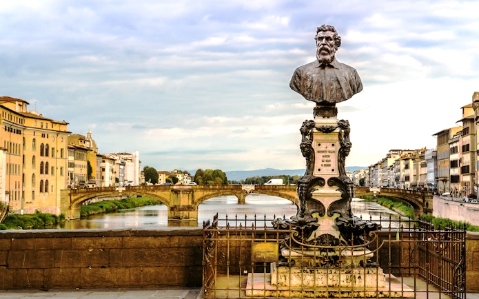 Bust of Benvenuto Cellini on Ponte Vecchio, Florence, with Arno River and historic buildings in view.