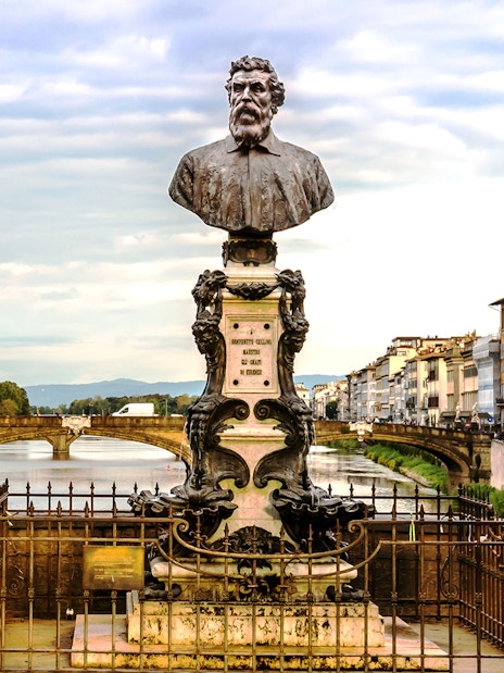 Bust of Benvenuto Cellini on Ponte Vecchio, Florence, with Arno River and historic buildings in view.