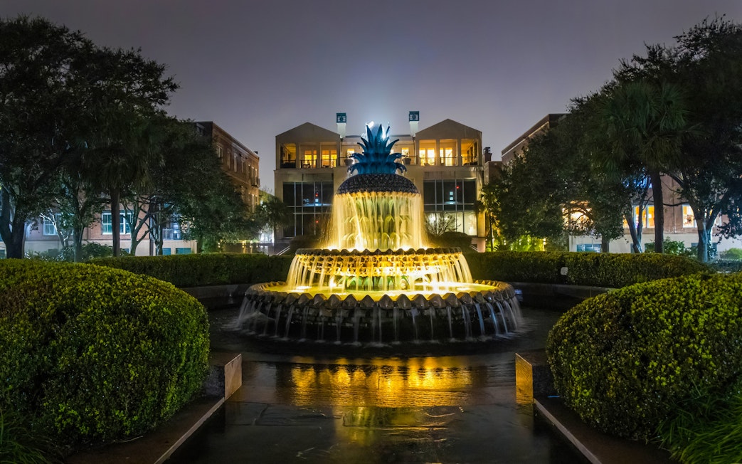 Illuminated Pineapple Fountain at night in Charleston, surrounded by trees and buildings.