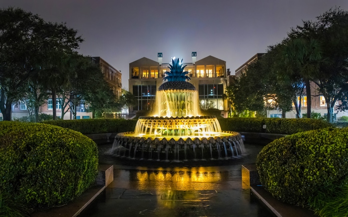 Illuminated Pineapple Fountain at night in Charleston, surrounded by trees and buildings.
