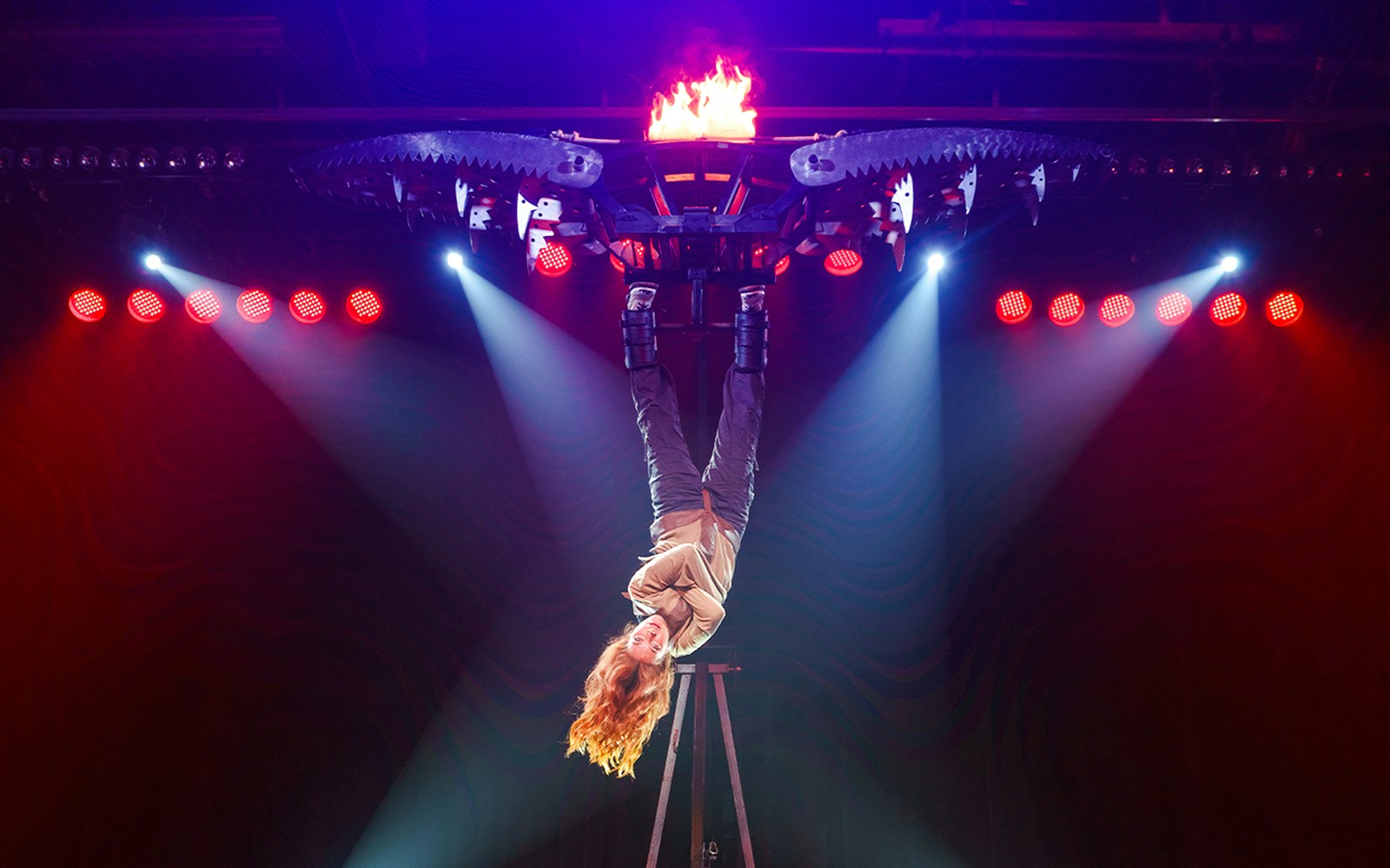 Performer suspended upside down with fire and saw blades at The Wild Magic Show, Greg Frewin Theatre.
