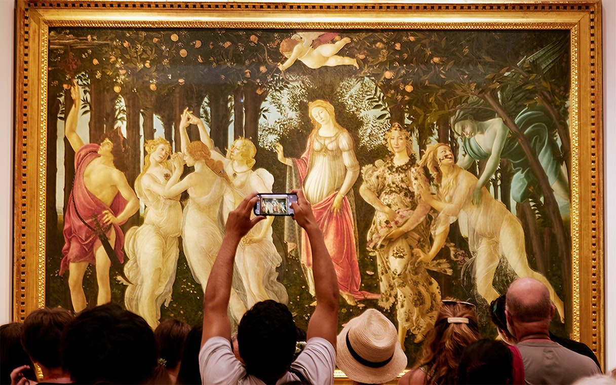 Tourists admire Botticelli's 'Primavera' at the Uffizi Gallery, with one capturing it on a smartphone.
