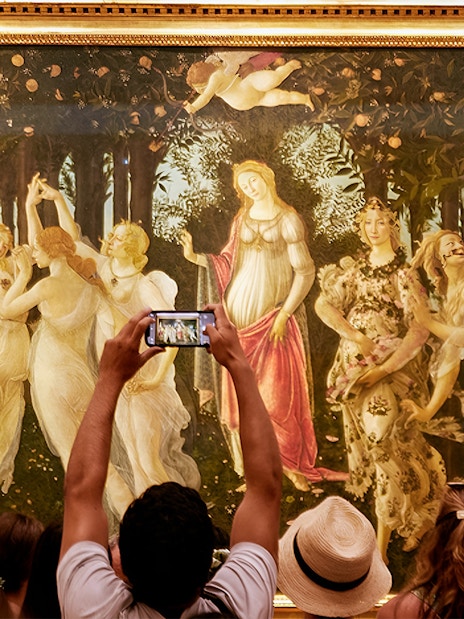 Tourists admire Botticelli's 'Primavera' at the Uffizi Gallery, with one capturing it on a smartphone.