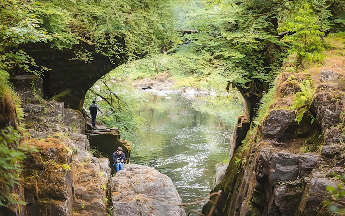 Visitors exploring a lush green gorge with a river on the Whisky & Waterfalls Day Tour from Edinburgh.