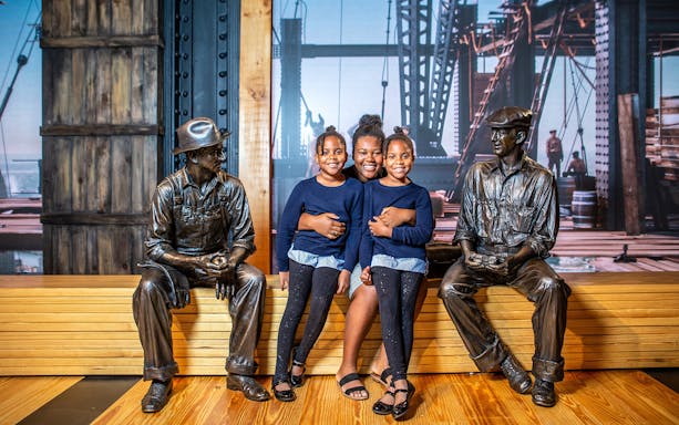 Visitors sitting with construction worker statues at Empire State Building exhibit.
