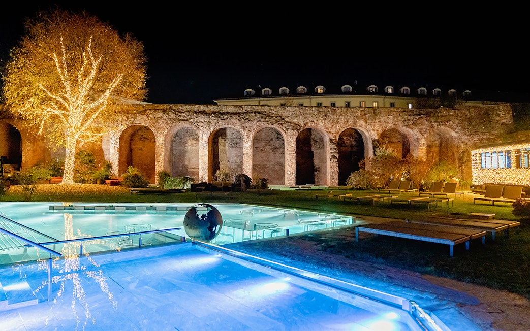 QC Terme Milano Resort & Spa outdoor pool with illuminated tree and ancient arches at night.