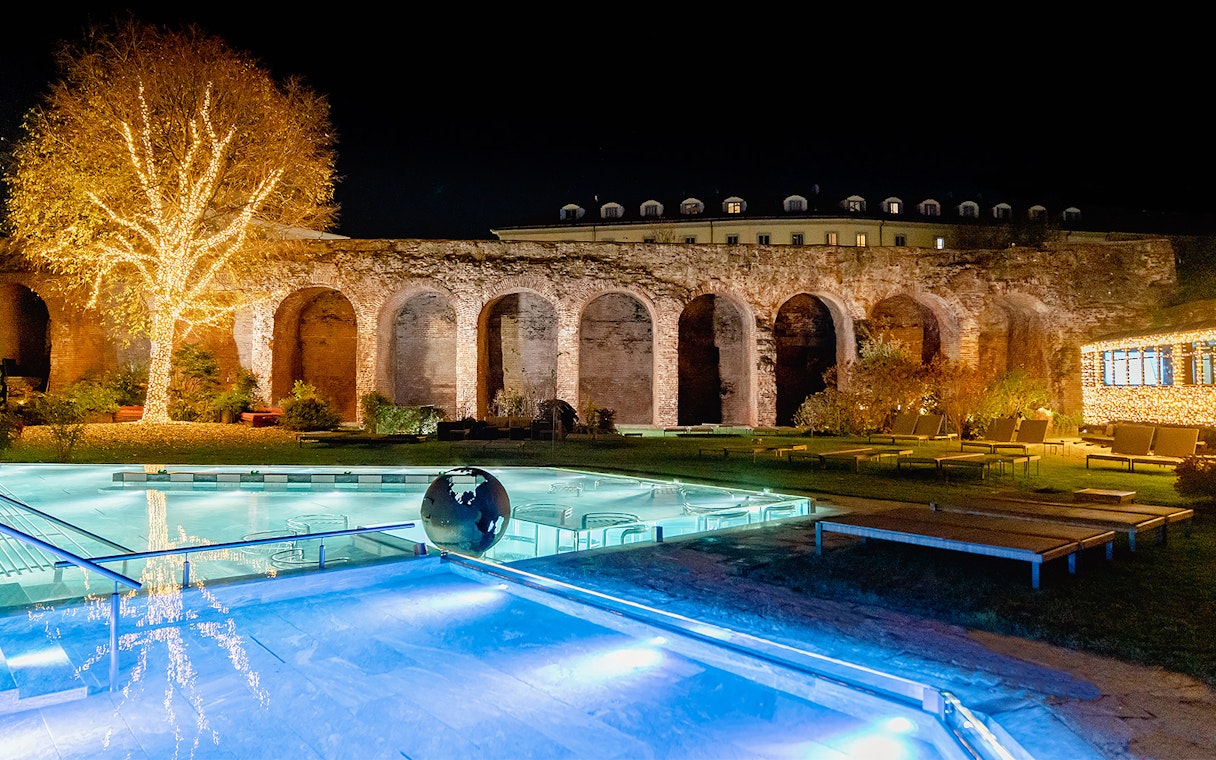 QC Terme Milano Resort & Spa outdoor pool with illuminated tree and ancient arches at night.