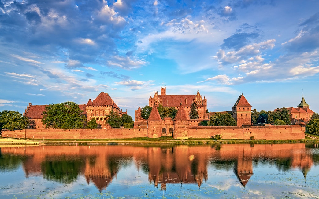 Malbork Castle reflected in the Nogat River under a blue sky.