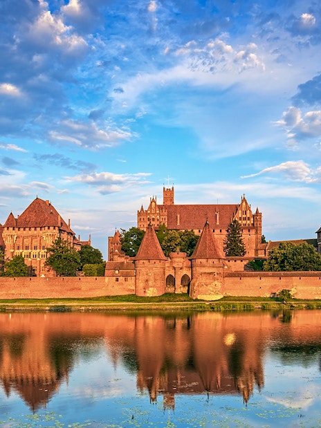 Malbork Castle reflected in the Nogat River under a blue sky.