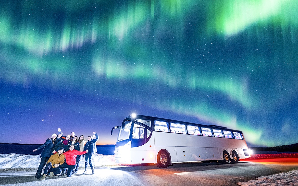 Tourists near a bus under Northern Lights in snowy landscape.