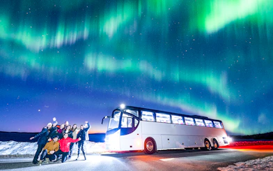 Tourists near a bus under Northern Lights in snowy landscape.