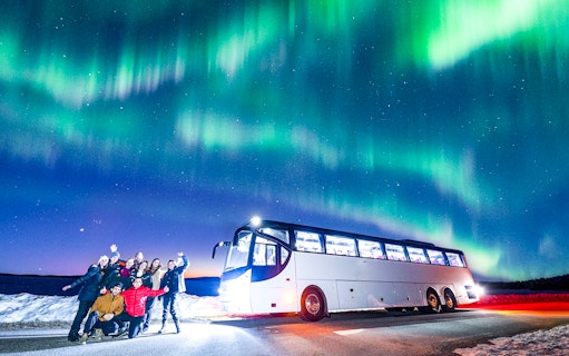 Tourists near a bus under Northern Lights in snowy landscape.