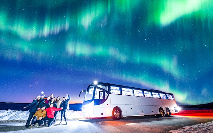 Tourists near a bus under Northern Lights in snowy landscape.