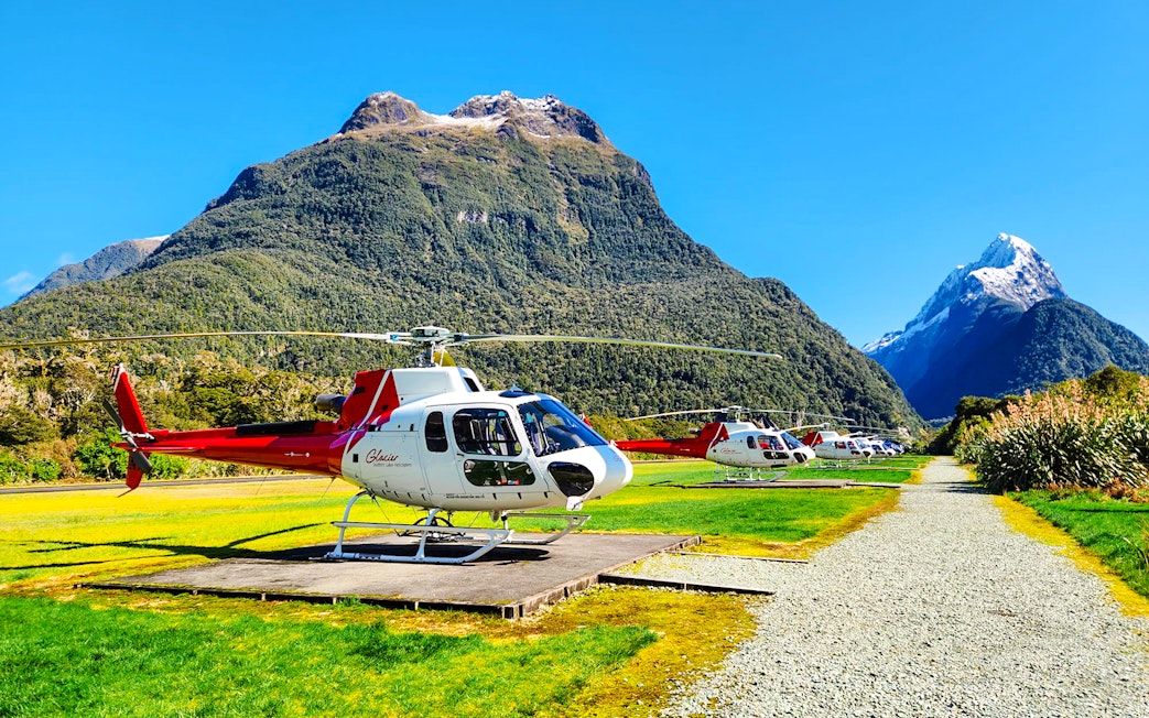 Helicopters on landing pads in a grassy field with Milford Sound mountains in the background.