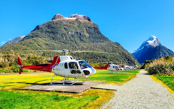 Helicopters on landing pads in a grassy field with Milford Sound mountains in the background.