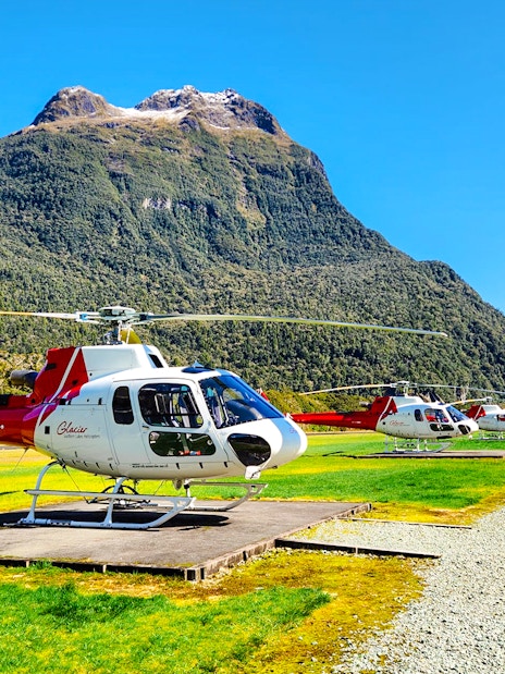 Helicopters on landing pads in a grassy field with Milford Sound mountains in the background.