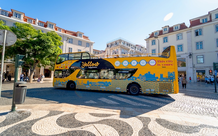 Yellow bus on Lisbon street during hop-on hop-off tour with historic buildings in background.