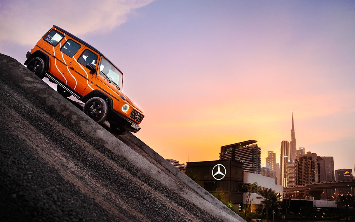 Mercedes-Benz G-Class on steep incline with Dubai skyline at sunset.