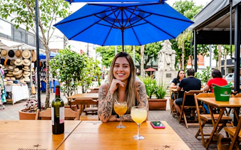 Woman enjoying drinks at an outdoor café in Rio de Janeiro, with a market and statue nearby.