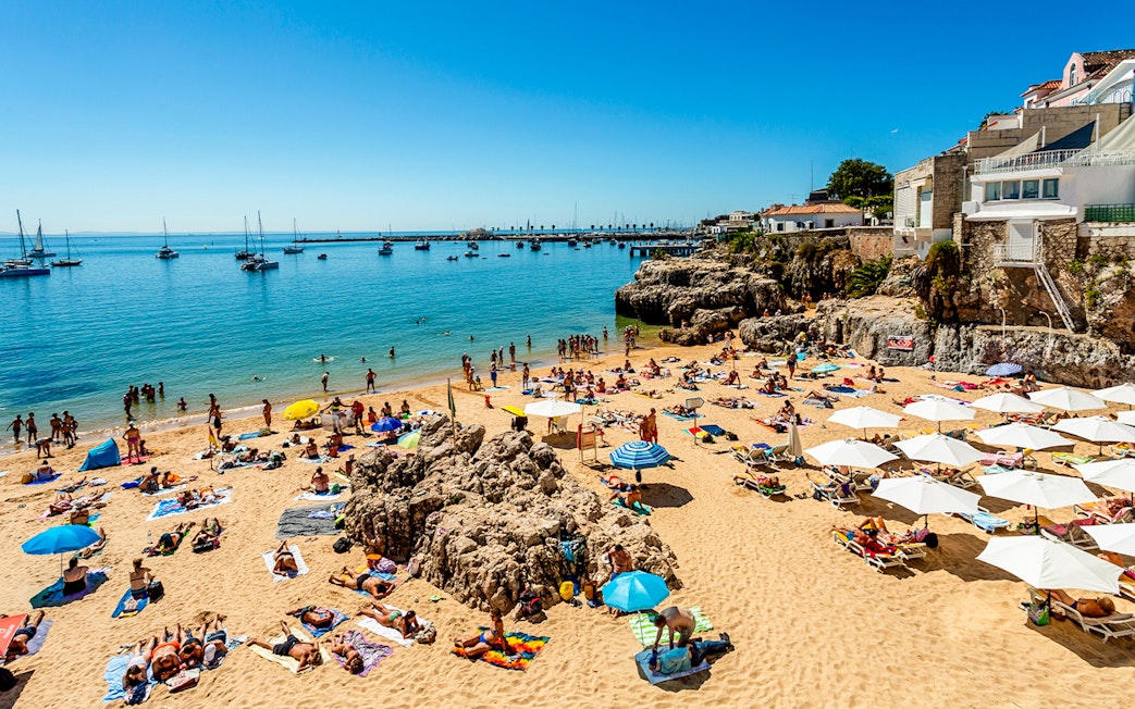 Beachgoers relaxing on Praia da Rainha in Cascais, Portugal, with boats in the distance.