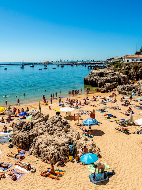 Beachgoers relaxing on Praia da Rainha in Cascais, Portugal, with boats in the distance.