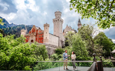 Tourists standing in front of Neuschwanstein Castle, Bavaria, surrounded by lush greenery.