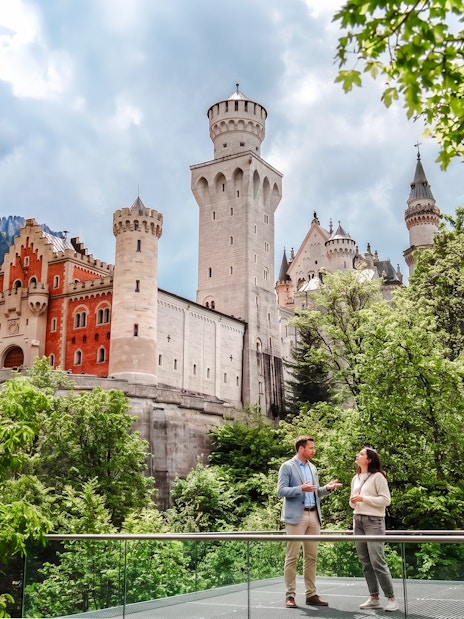 Tourists standing in front of Neuschwanstein Castle, Bavaria, surrounded by lush greenery.
