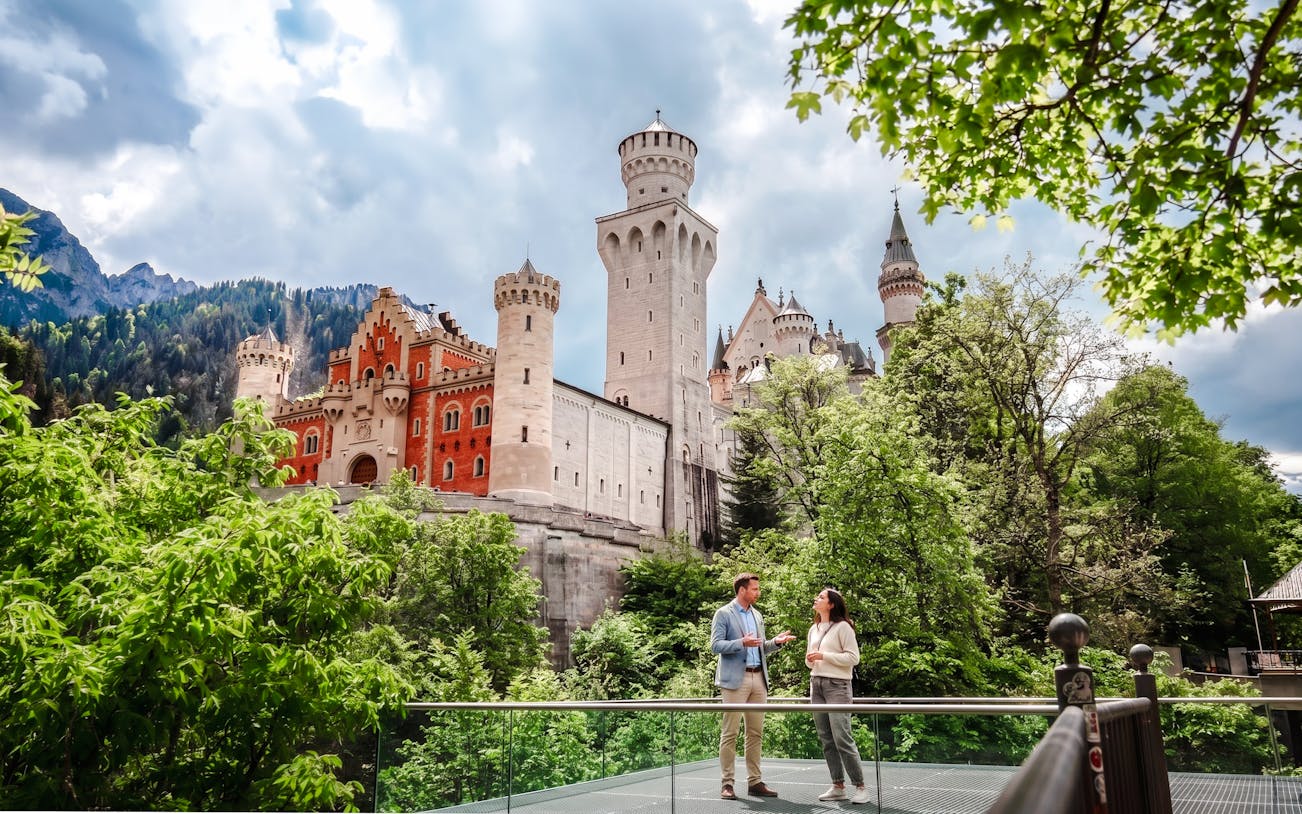 Tourists standing in front of Neuschwanstein Castle, Bavaria, surrounded by lush greenery.