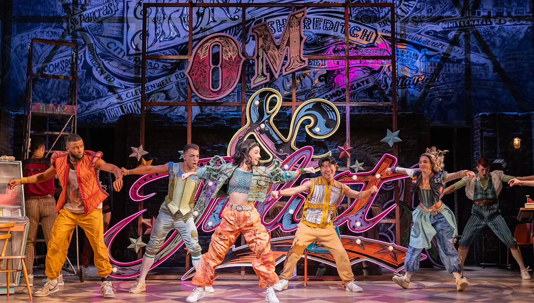 Performers dancing energetically on stage in the musical "& Juliet" with vibrant neon backdrop.