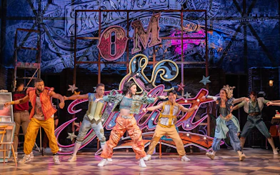 Performers dancing energetically on stage in the musical "& Juliet" with vibrant neon backdrop.