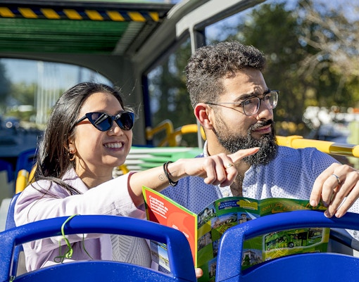 Couple sitting on the upper deck of a blue bus, holding a city pamphlet n one hand and enjoying their trip
