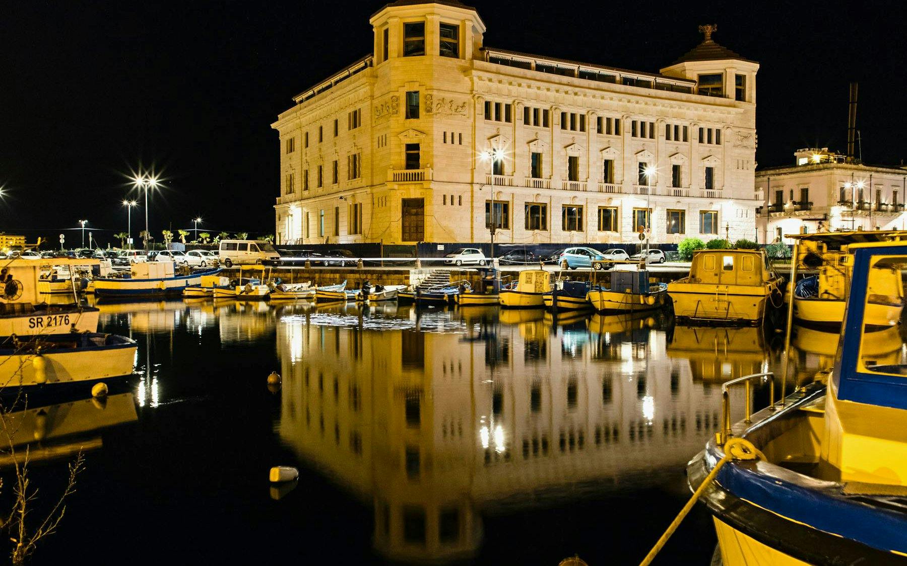 Ortigia waterfront at night with boats and illuminated historic building.