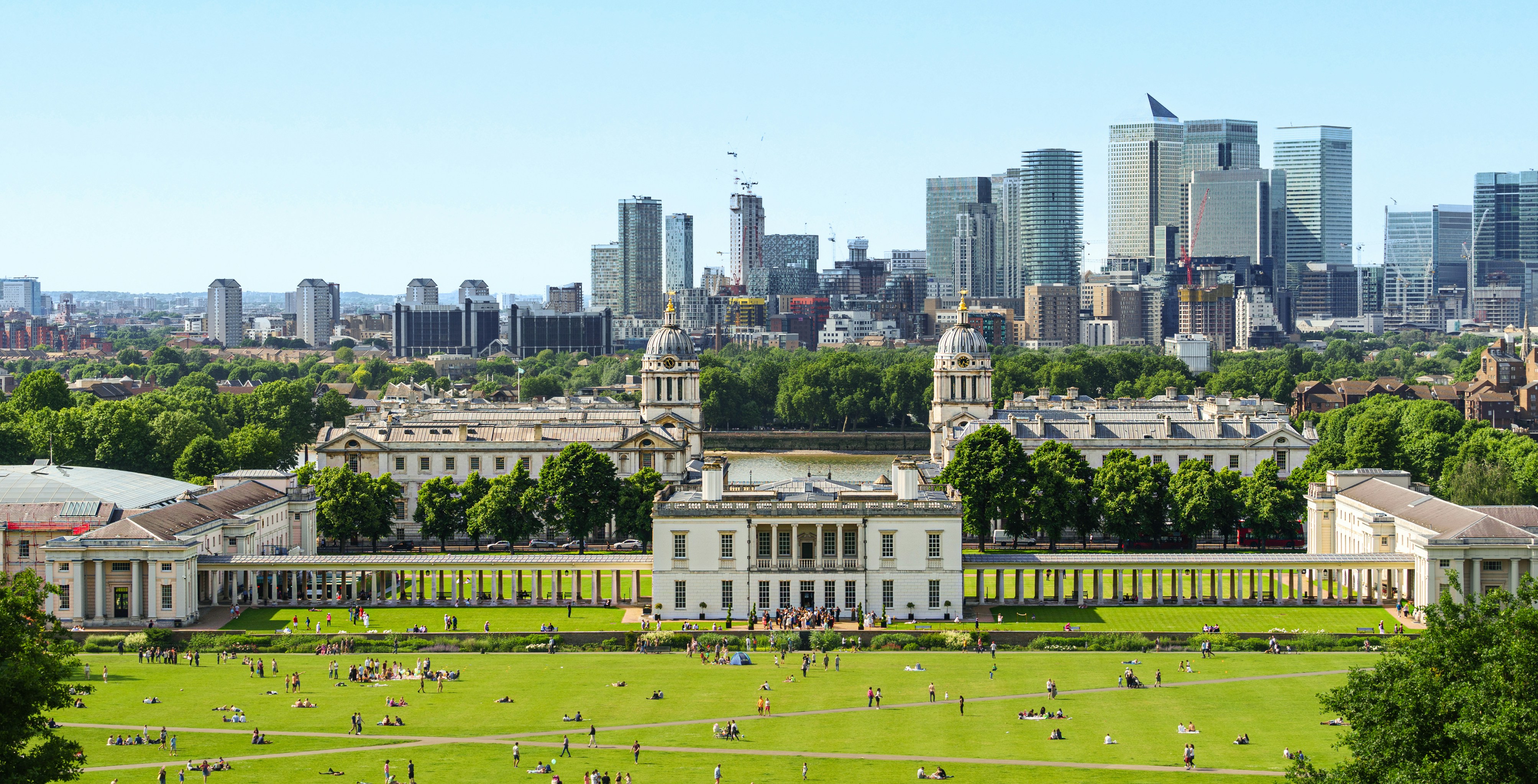 Panoramic view of Canary Wharf skyline from Greenwich Park, London, with historic buildings in foreground.