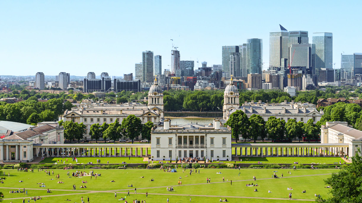 Panoramic view of Canary Wharf skyline from Greenwich Park, London, with historic buildings in foreground.