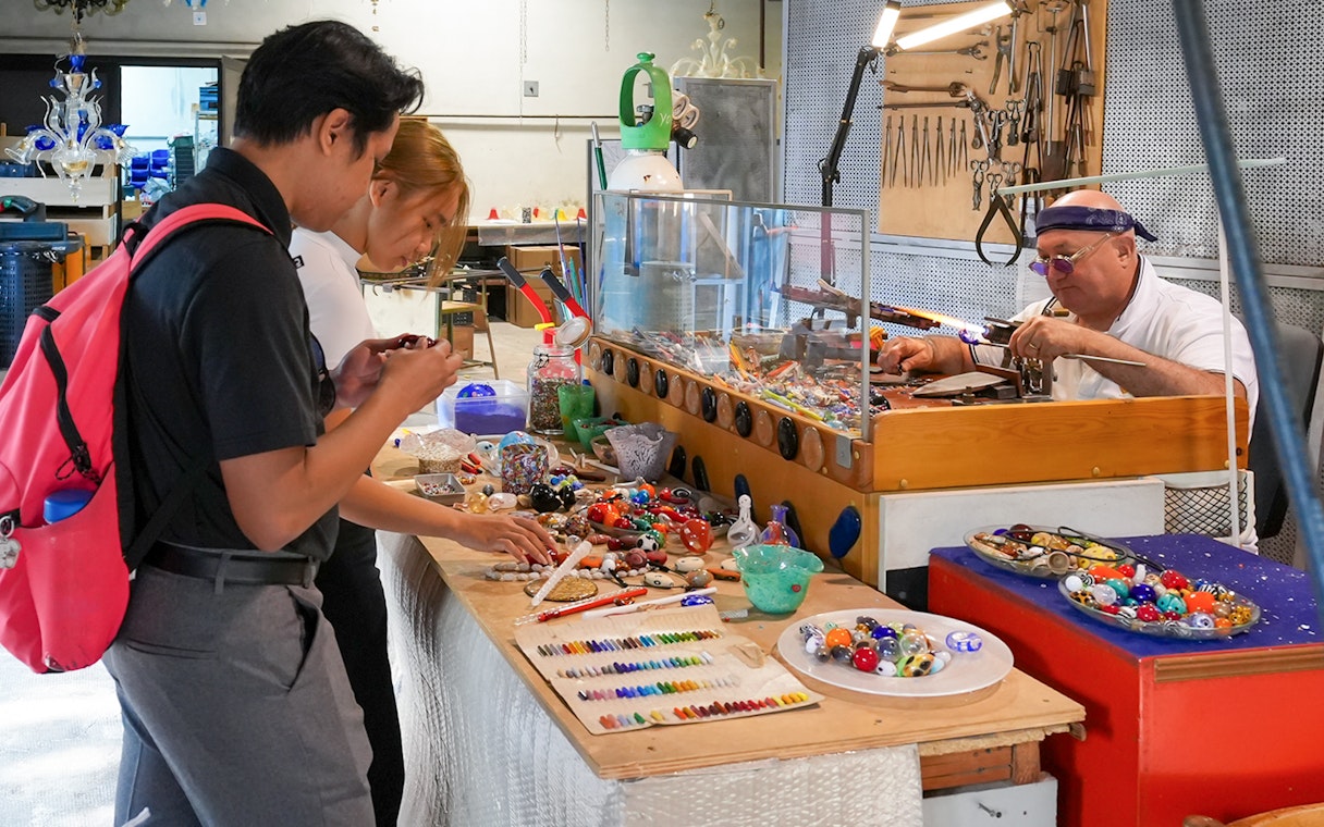 Glass artist crafting intricate pieces in a showroom as visitors observe.