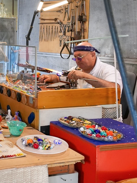 Glass artist crafting intricate pieces in a showroom as visitors observe.