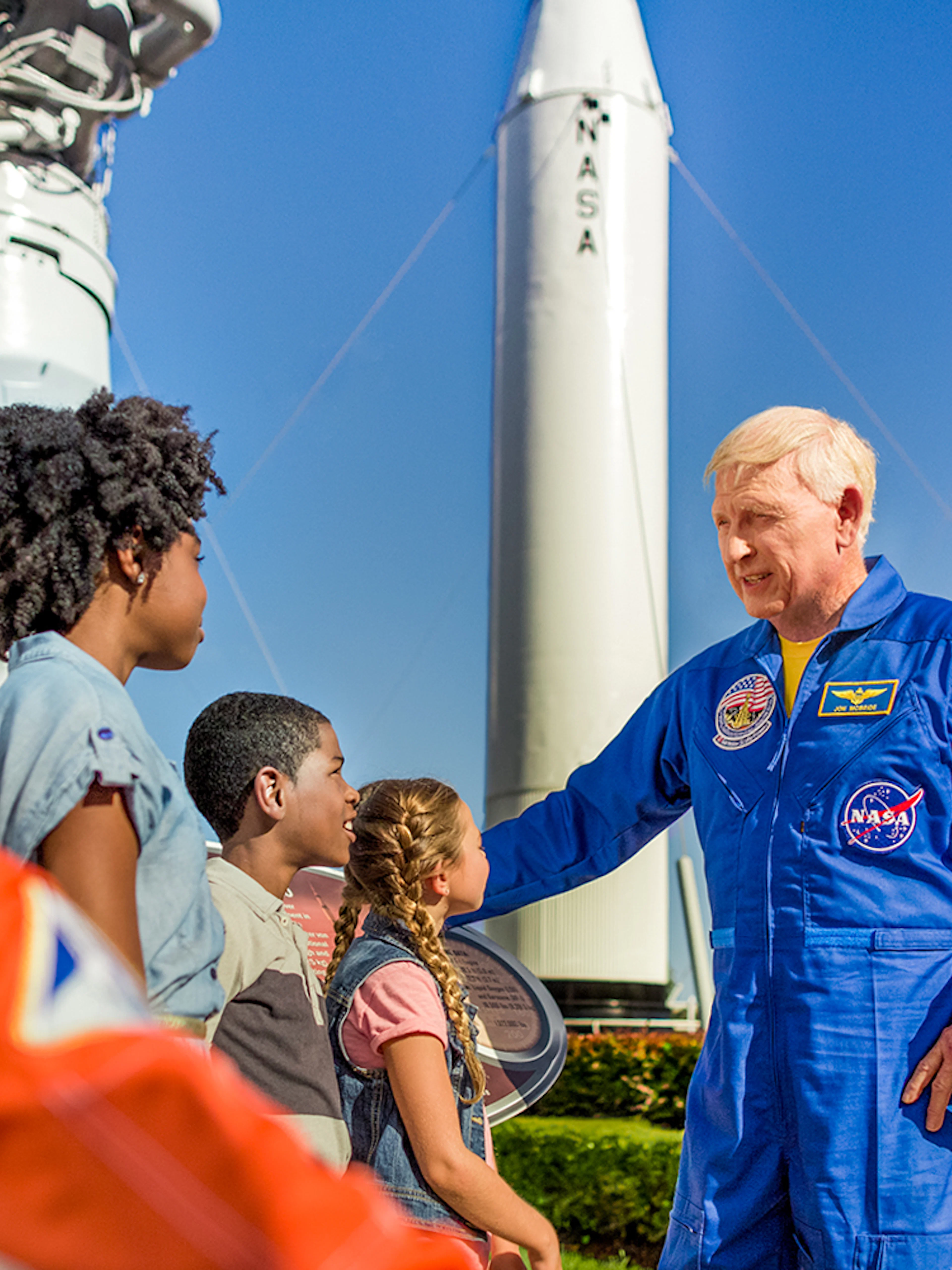 Astronaut interacting with children at Kennedy Space Center, rockets in background.
