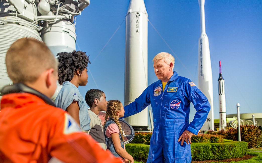Astronaut interacting with children at Kennedy Space Center, rockets in background.