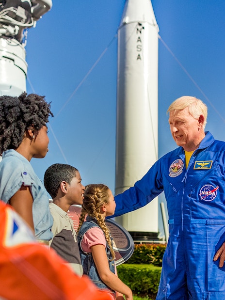 Astronaut interacting with children at Kennedy Space Center, rockets in background.