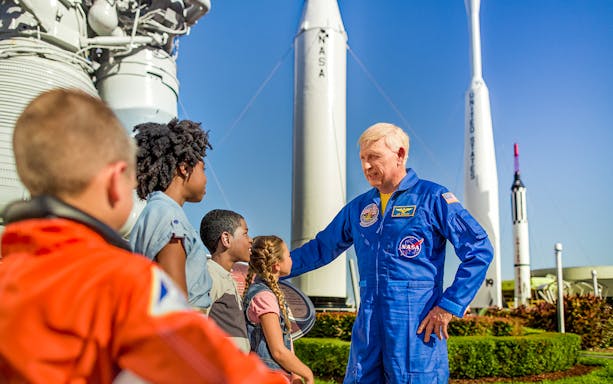Astronaut interacting with children at Kennedy Space Center, rockets in background.