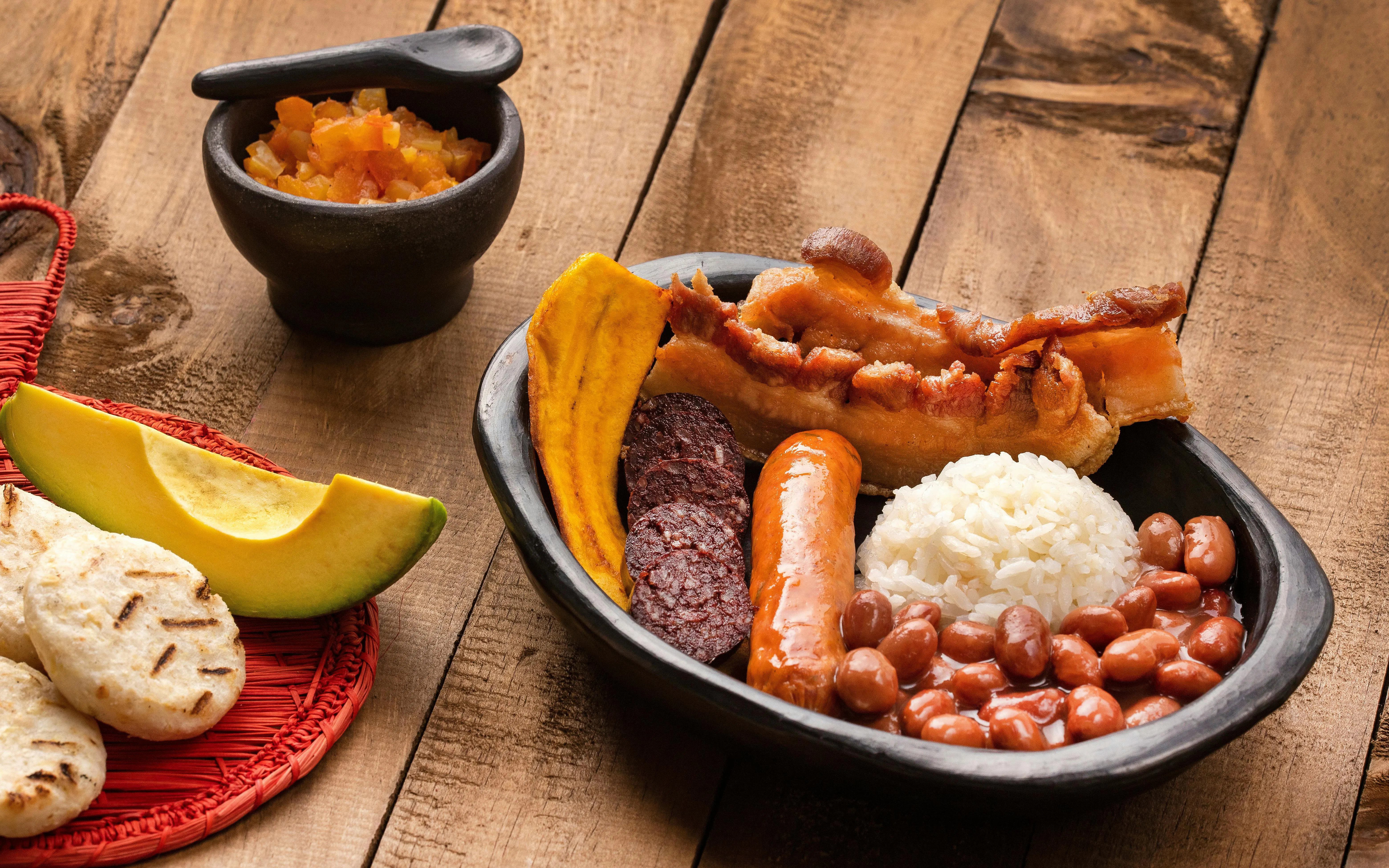 Bandeja Paisa with rice, beans, chorizo, chicharrón, plantain, avocado, and arepas on a wooden table.