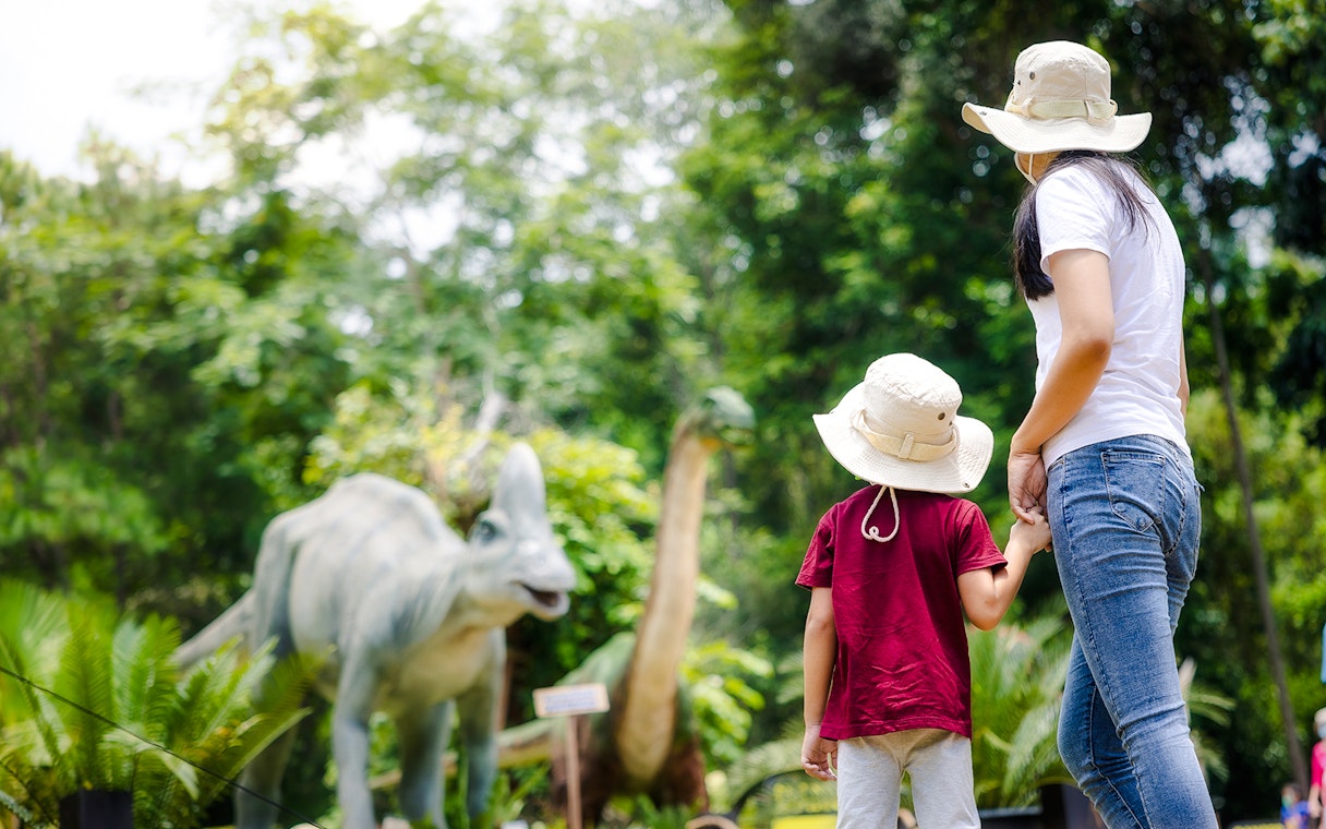 Mom and child viewing dinosaur models in a lush park setting.