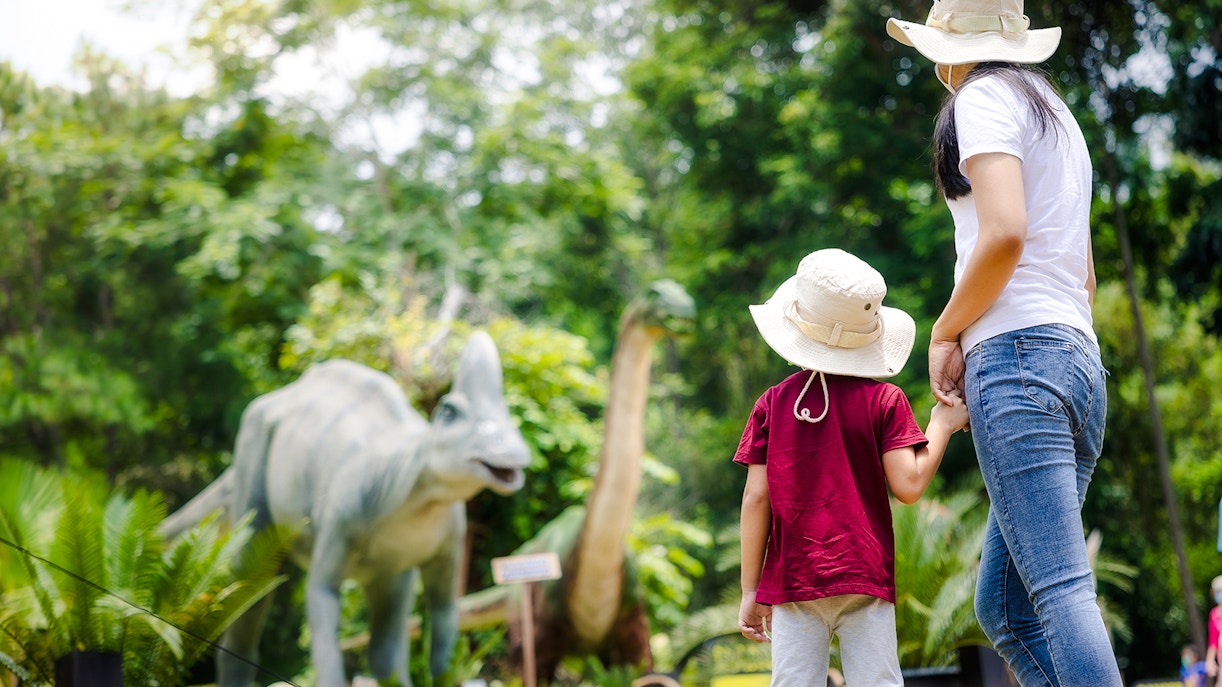 Mom and child viewing dinosaur models in a lush park setting.
